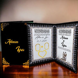 A black and gold Nikah certificate book with intricate gold corner embellishments, customized with the names "Adnan & Izra" in gold script. The interior features Quranic calligraphy, thumbprint spaces, and a section for signatures.
