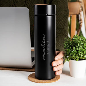 Hand holding a personalized black thermos with custom engraving, placed next to a laptop and potted plant.