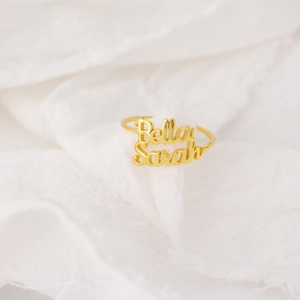 Custom name ring in gold with the names "Bella" and "Sarah" engraved, displayed on a white fabric background