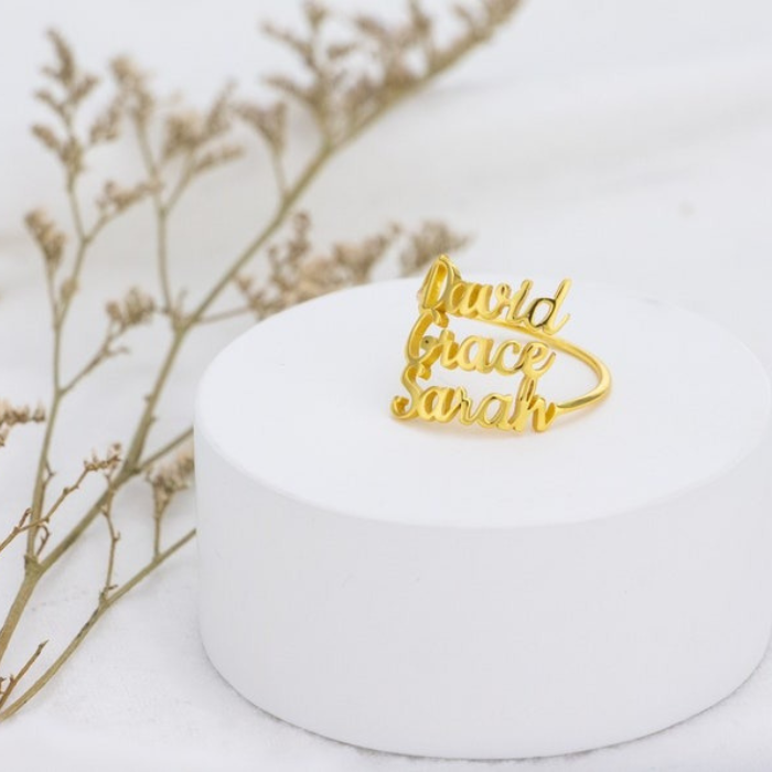 Custom name ring in gold with the names "David," "Grace," and "Sarah" engraved, displayed on a white stand with dried flowers in the background