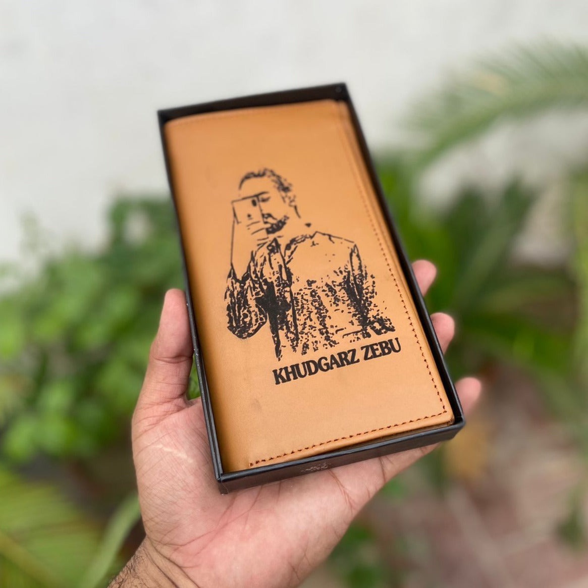 Custom picture wallet with photo and name "Khudgarz Zebu" on tan leather, held in hand with a black gift box and green plants in the background