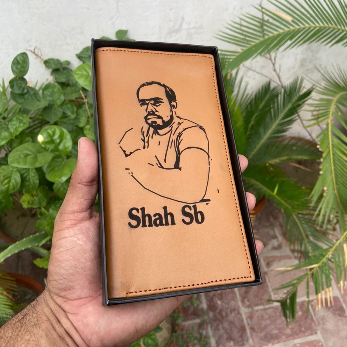 Custom picture wallet with photo and name "Shah SB" on tan leather, held in hand with a black gift box and green plants in the background