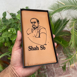 Custom picture wallet with photo and name "Shah SB" on tan leather, held in hand with a black gift box and green plants in the background