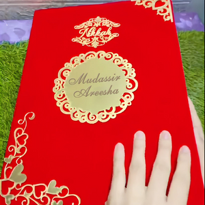 Customized velvet Nikkah Nama certificate with "Mudassir & Areesha" text, featuring gold heart accents on a red velvet cover, held by a hand against a green backdrop