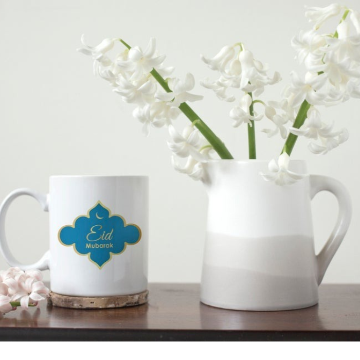 Eid Mubarak Mug with blue and gold design, placed next to a white pitcher of flowers on a wooden surface.