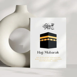 A modern Hajj Mubarak greeting card placed against a minimalist ceramic vase, featuring a Kaaba illustration and a Quranic blessing. A heartfelt Islamic gift for loved ones.
