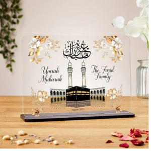 A beautifully designed Umrah Mubarak acrylic plaque featuring a Kaaba illustration, Arabic calligraphy, and customizable text, placed on a wooden base with floral decorations.