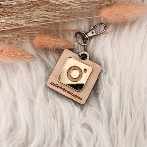 Personalized NFC keychain featuring a golden Instagram emblem on a wooden backing, engraved with a custom handle, placed on a soft fur background with decorative dried grass accents.