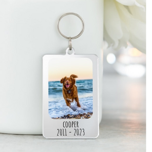 A custom pet memorial keychain featuring a photo of a dog playing on the beach with engraved text "Cooper 2011 - 2023."