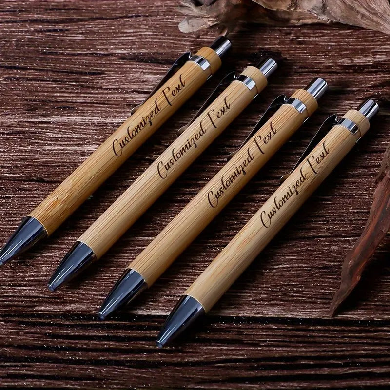 A set of four customized bamboo wood ballpoint pens with engraved text, placed on a rustic wooden surface.