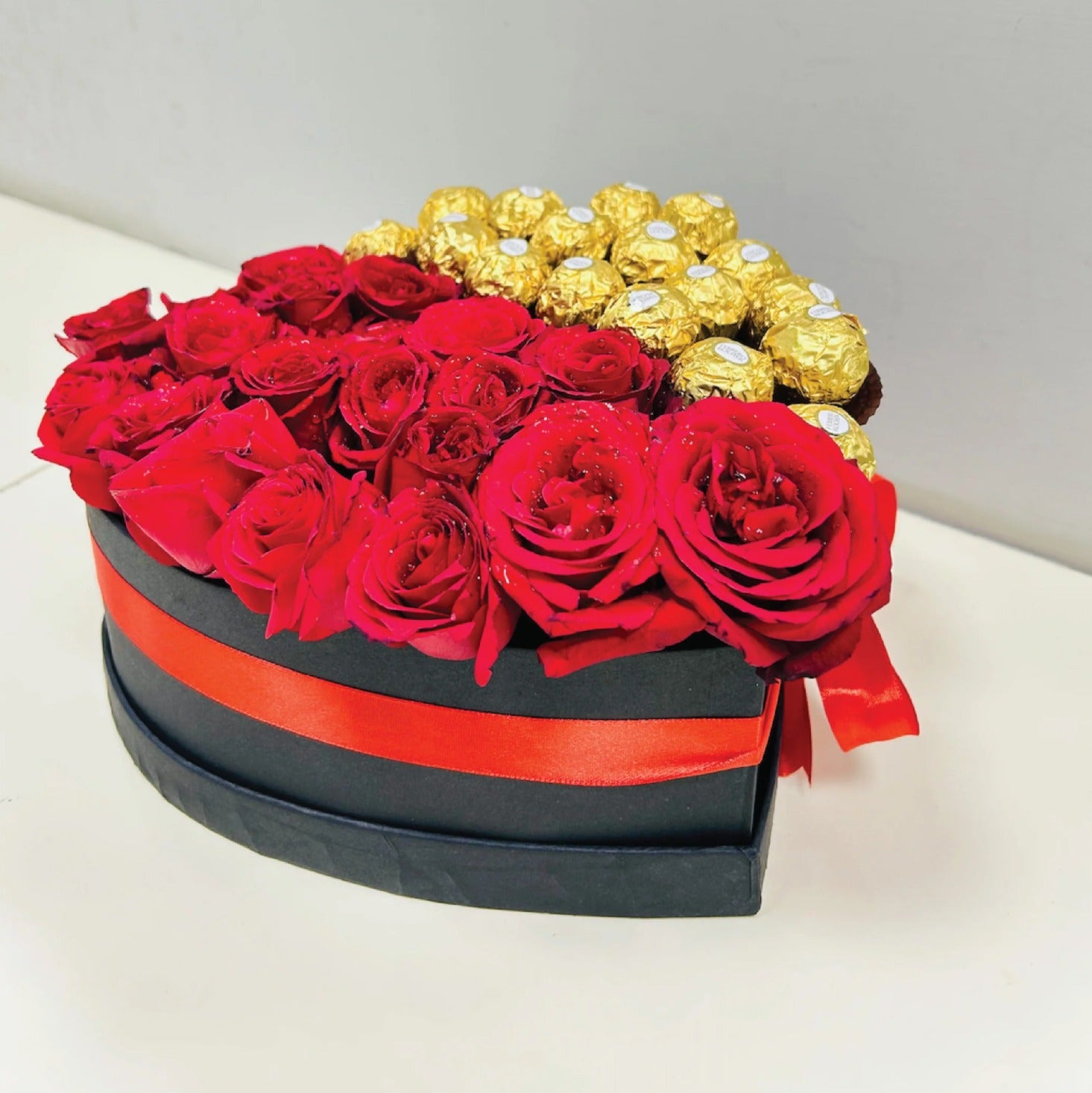 Elegant side view of a heart-shaped luxury gift box with Ferrero Rocher chocolates and red roses, tied with a red satin ribbon, perfect for gifting.