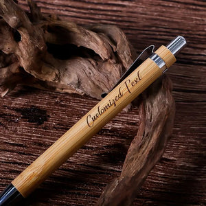 A close-up of a single customized bamboo wood ballpoint pen with engraved text, resting on a natural wooden surface.