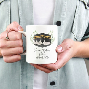 A person holding a personalized Umrah Mubarak mug featuring the Kaaba design, custom name, and year 2024/1445. The mug is a meaningful Islamic gift for returning Umrah pilgrims.