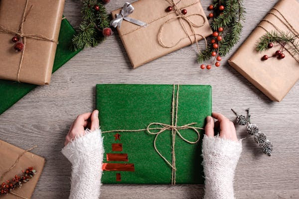 Hands holding green gift box with rustic twine bow, surrounded by holiday wrapped presents and festive greenery