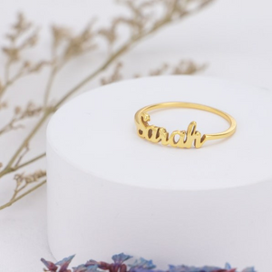 Custom name ring in gold with the names "Sarah"engraved, displayed on a white stand with dried flowers in the background