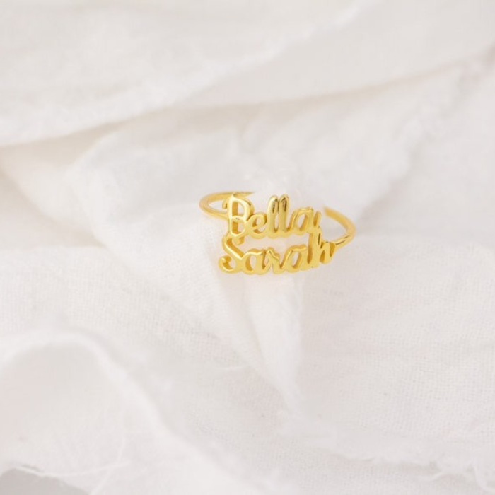 Custom name ring in gold with the names "Bella" and "Sarah" engraved, displayed on a white fabric background