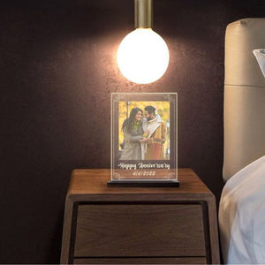 Personalized anniversary night lamp with "Happy Anniversary 4/16/2022" message on a wooden bedside table, softly lit by a hanging bulb in a cozy bedroom setting