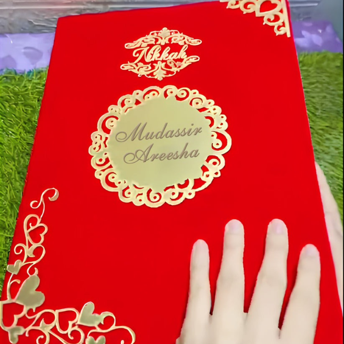 Customized velvet Nikkah Nama certificate with "Mudassir & Areesha" text, featuring gold heart accents on a red velvet cover, held by a hand against a green backdrop