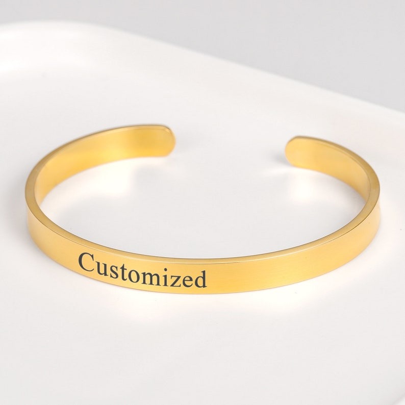 Engraved cuff bangle bracelet in gold with the word "Customized," displayed on a white surface