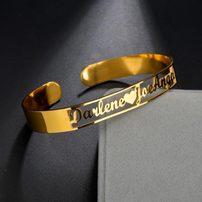 Engraved cuff bangle bracelet in gold with the names "Darlene" and "Joe Angel" and a heart accent, displayed on a gray surface