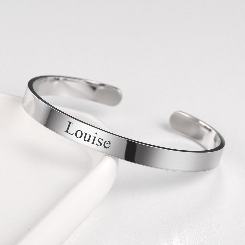Engraved cuff bangle bracelet in silver with the name "Louise," displayed on a white surface