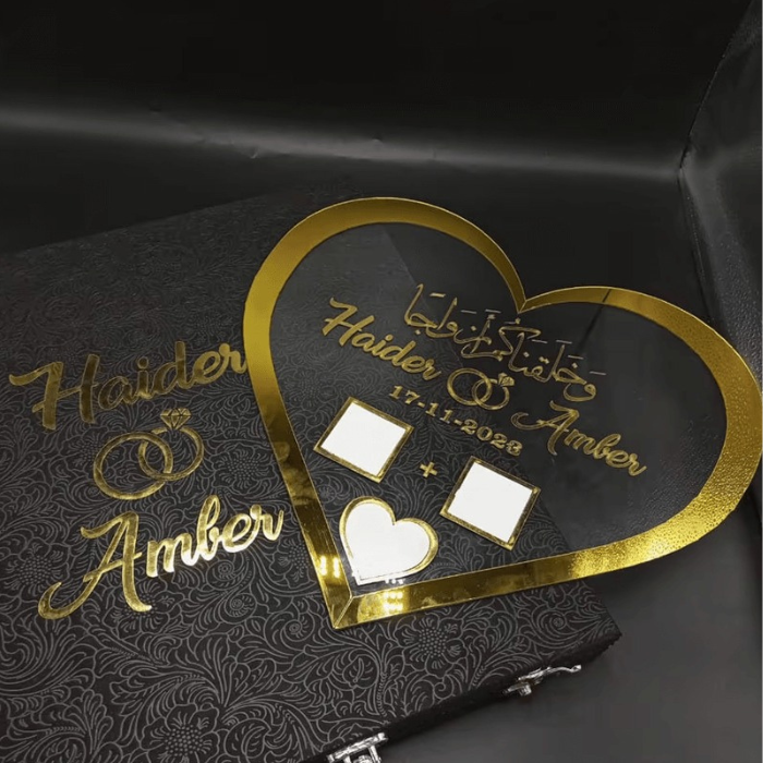 Heart-shaped Nikah thumbprint acrylic board personalized with "Haider & Amber" and "17-11-2028," showcasing a gold heart frame and thumbprint areas, placed on a black velvet book