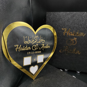 Heart shaped Nikah thumb acrylic board with "Haider & Amber" and "17-11-2028" text, featuring gold heart border and thumbprint spaces, on a black velvet book