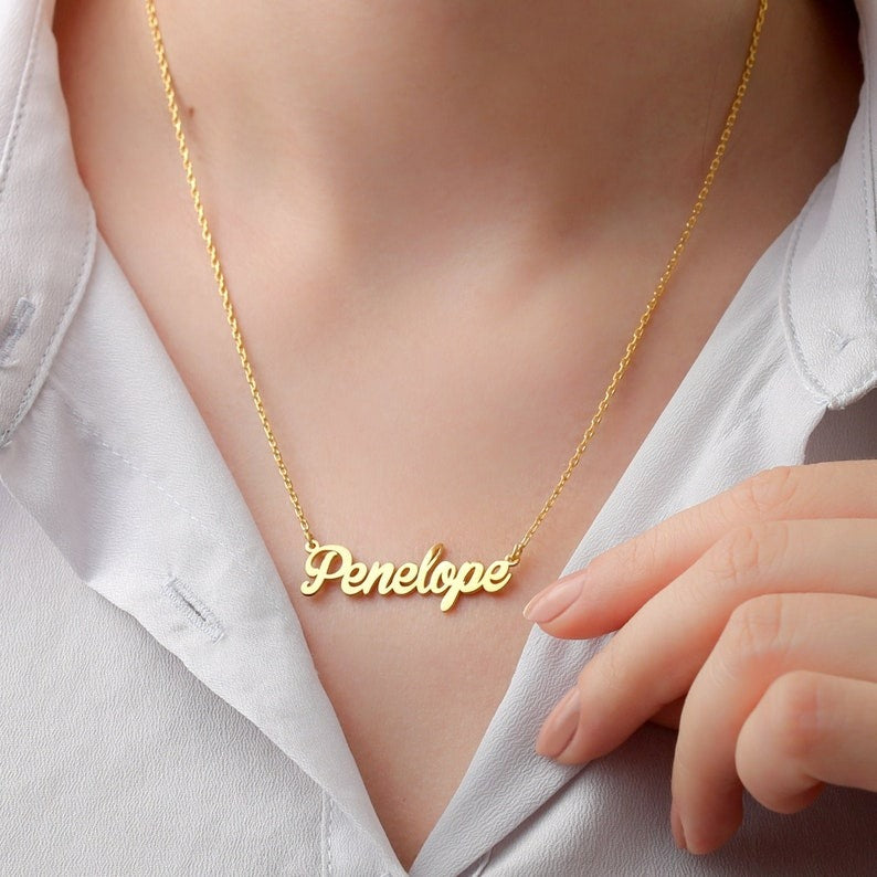 Personalized jewelry name necklace in gold with the name "Penelope," worn on a white shirt, held by hand