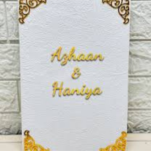 Custom Nikkah Nama velvet booklet with "Azhaan & Haniya" in gold foil, featuring elegant corner designs, placed against a white brick wall background
