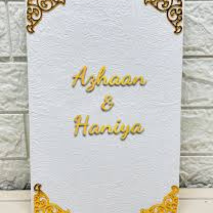 Custom Nikkah Nama velvet booklet with "Azhaan & Haniya" in gold foil, featuring elegant corner designs, placed against a white brick wall background