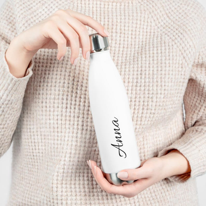 : A woman holding a sleek, white insulated stainless steel water bottle with the name "Anna" elegantly engraved in black script. The bottle features a secure, leak-proof stainless steel cap, ideal for keeping beverages hot or cold for extended hours.