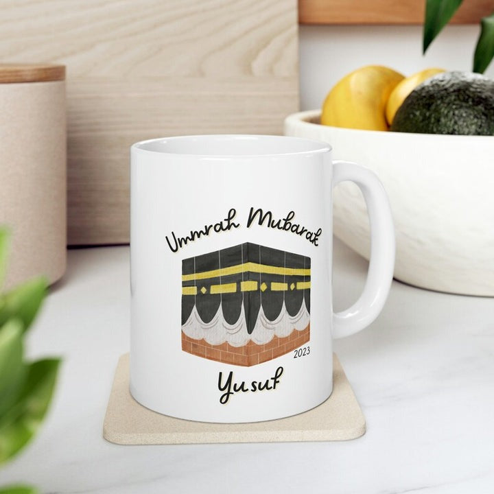 A white ceramic mug featuring an illustration of the Kaaba, with the text "Umrah Mubarak" and the name "Yusuf" in stylish fonts, symbolizing a personalized Umrah gift.