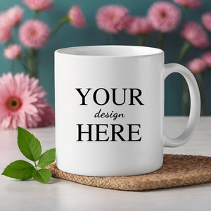 White ceramic mug featuring 'Your Design Here' text placed on a woven coaster, with pink flowers and green leaves in the background.