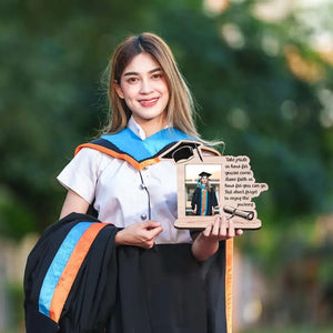 Graduate in a cap and gown holding a diploma and wooden graduation photo frame with "Take Pride" quote, displayed indoors with a festive background