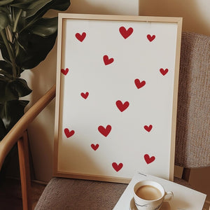 Framed minimalist red hearts wall art resting on a chair beside a coffee cup, creating a cozy and romantic home decor aesthetic.