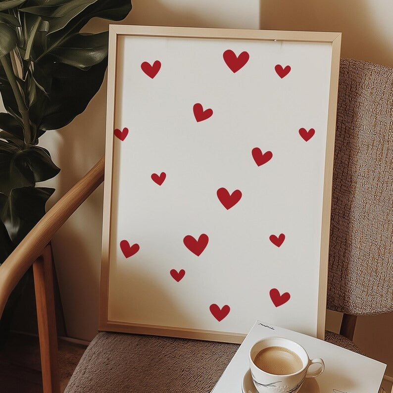 Framed minimalist red hearts wall art resting on a chair beside a coffee cup, creating a cozy and romantic home decor aesthetic.