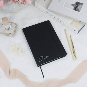 Elegant black hardback notebook with a personalized name, surrounded by stationery items on a white table.