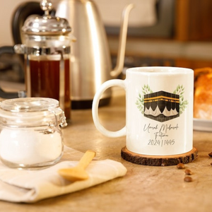 A personalized white ceramic mug featuring an Umrah Mubarak design with an illustration of the Kaaba, surrounded by green leaves. The mug includes a custom name and the year 2024/1445, making it a thoughtful gift for Umrah travelers.