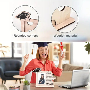 Graduate holding a wooden graduation photo frame with "Take Pride" quote and cap design, displayed indoors with a laptop and decor in the background